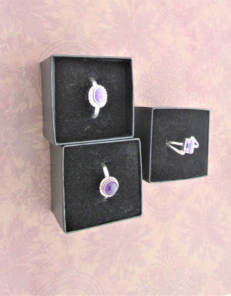 May include: Three small black boxes with velvet lining, each containing a silver ring with a purple gemstone.