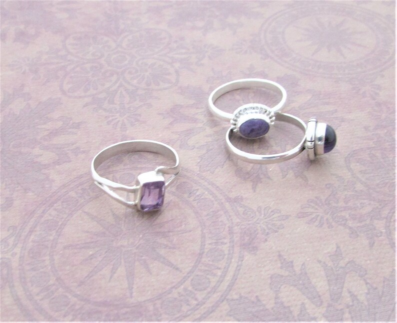 May include: Two silver rings with purple amethyst gemstones. One ring has a round stone and the other has a rectangular stone.