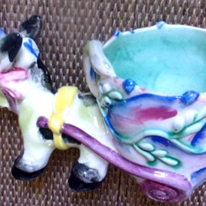 Peut inclure: Une figurine en céramique représentant un âne tirant une charrette. L'âne est blanc avec des taches noires, un harnais jaune et un nœud rose. La charrette est peinte de motifs floraux bleus, roses et verts, avec un intérieur vert clair et une roue violette.