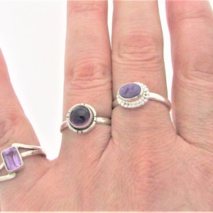 May include: Three silver rings with purple amethyst gemstones. The rings are on a person's hand.