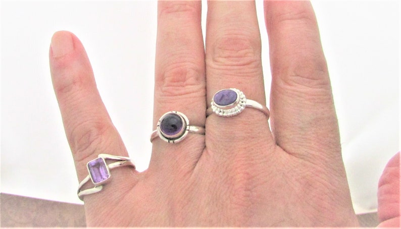 May include: Three silver rings with purple amethyst gemstones. The rings are on a person's hand.
