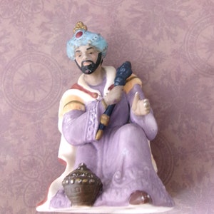 Puede incluir: Una figurita de porcelana de uno de los tres reyes magos, vestido con una túnica morada con ribetes dorados, un turbante azul y sosteniendo un bastón. Está sentado en un trono y sostiene una caja de oro.