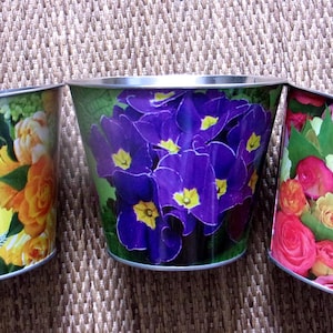 Może przedstawiać: Trzy metalowe doniczki z kwiatowymi wzorami. Jedna doniczka ma żółte i pomarańczowe kwiaty, druga fioletowe, a trzecia różowe i żółte róże. Każda doniczka ma srebrną obwódkę.