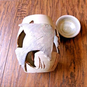 May include: A beige ceramic candle holder with a cutout design featuring a large fish relief on the front. The cylindrical vessel has a textured, matte finish with dark brown accents along the edges of the cutouts and includes a small matching lid.