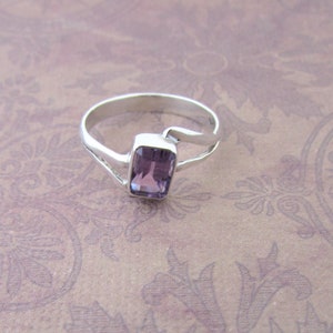 May include: A silver ring with a purple amethyst gemstone set in a rectangular shape. The ring has a unique, curved design.