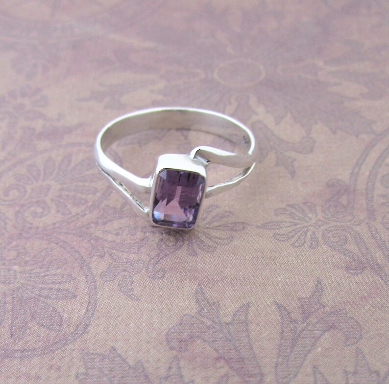 May include: A silver ring with a purple amethyst gemstone set in a rectangular shape. The ring has a unique, curved design.