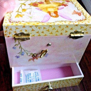K&ouml;nnte beinhalten: Eine rosa-gelbe Spieluhr mit einer Feenillustration auf dem Deckel. Die Box hat ein Blumenmuster und goldfarbene Beschl&auml;ge. Die Schublade ist ge&ouml;ffnet und zeigt einen rosa Innenraum und ein Trockenmittelbeutel.