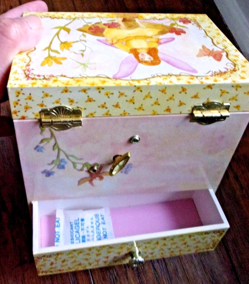 K&ouml;nnte beinhalten: Eine rosa-gelbe Spieluhr mit einer Feenillustration auf dem Deckel. Die Box hat ein Blumenmuster und goldfarbene Beschl&auml;ge. Die Schublade ist ge&ouml;ffnet und zeigt einen rosa Innenraum und ein Trockenmittelbeutel.