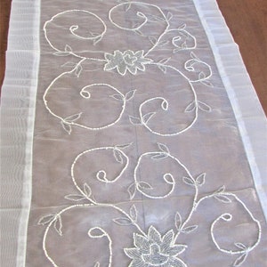 May include: White beaded table runner with a floral design. The runner is made of sheer fabric and has a delicate, feminine look.