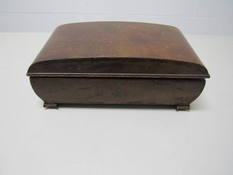 Puede incluir: Una caja de madera vintage con tapa curvada y acabado marr&oacute;n oscuro. La caja tiene forma rectangular con bordes redondeados y se apoya sobre peque&ntilde;os pies. La caja probablemente se utiliza para el almacenamiento.