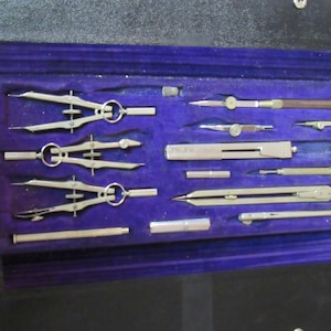 May include: A vintage set of drafting tools in a black leather case with a purple velvet lining. The tools include compasses, rulers, and a protractor.