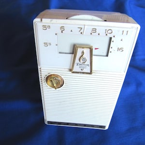May include: A vintage white Emerson radio with a gold dial and tuning dial. The rectangular radio has horizontal lines on the front. The Emerson logo is displayed on the front. The radio has a retro design.