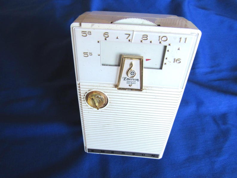 May include: A vintage white Emerson radio with a gold dial and tuning dial. The rectangular radio has horizontal lines on the front. The Emerson logo is displayed on the front. The radio has a retro design.
