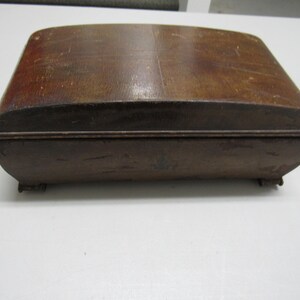 Puede incluir: Una caja rectangular de madera con tapa y base curvadas. La caja es de madera marr&oacute;n oscuro con vetas visibles y un aspecto ligeramente desgastado. La caja tiene peque&ntilde;os pies y un dise&ntilde;o simple y cl&aacute;sico.
