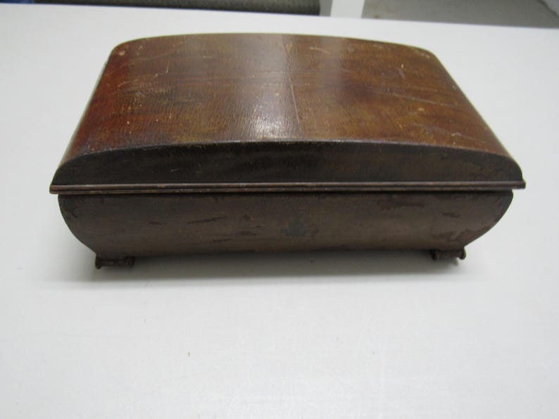 Puede incluir: Una caja rectangular de madera con tapa y base curvadas. La caja es de madera marr&oacute;n oscuro con vetas visibles y un aspecto ligeramente desgastado. La caja tiene peque&ntilde;os pies y un dise&ntilde;o simple y cl&aacute;sico.