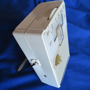 May include: A vintage white portable radio with a gold-coloured tuning knob and dial. The radio has a built-in stand and a clear dial with numbers. The radio is rectangular and has a ribbed front panel.