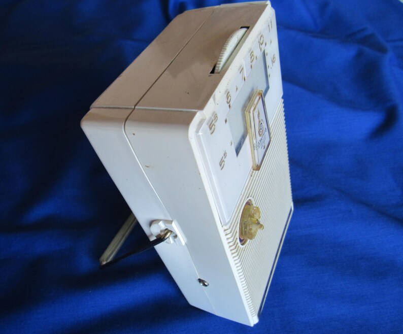 May include: A vintage white portable radio with a gold-coloured tuning knob and dial. The radio has a built-in stand and a clear dial with numbers. The radio is rectangular and has a ribbed front panel.