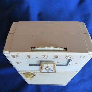 May include: An off-white vintage Emerson radio with a dial and speaker. The radio has a rectangular shape with a slightly angled front. The dial is a semi-circle shape. The radio has a gold-coloured emblem.
