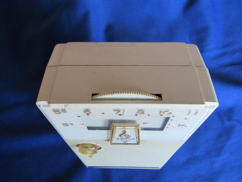 May include: An off-white vintage Emerson radio with a dial and speaker. The radio has a rectangular shape with a slightly angled front. The dial is a semi-circle shape. The radio has a gold-coloured emblem.
