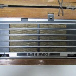 Puede incluir: Radio Emerson vintage con una caja de madera. La parte frontal presenta una rejilla de altavoz con barras plateadas horizontales y un panel negro. La palabra "EMERSON" se muestra en la parte delantera. La radio tiene una perilla de control de volumen.