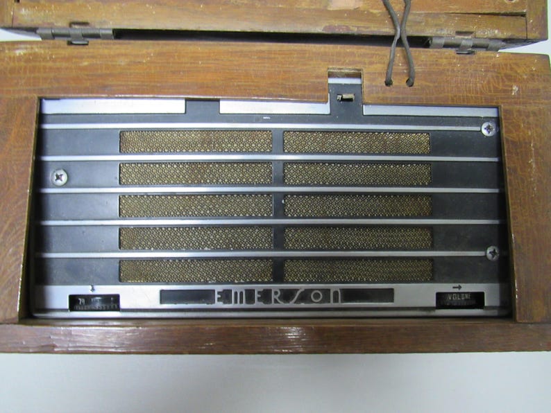 Puede incluir: Radio Emerson vintage con una caja de madera. La parte frontal presenta una rejilla de altavoz con barras plateadas horizontales y un panel negro. La palabra "EMERSON" se muestra en la parte delantera. La radio tiene una perilla de control de volumen.