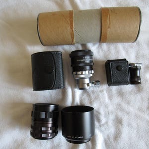 May include: A set of camera lenses and accessories, including a black lens cap, a black lens hood, a black leather case, a silver lens with the text "A.S.S.R.  MICROSCOPE  RANGE" and a black and silver lens with a black leather case.