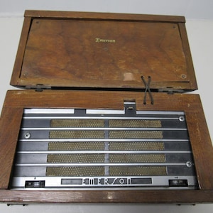 Puede incluir: Radio Emerson vintage con una caja de madera. La radio tiene una tapa con bisagras y una rejilla de altavoz con barras met&aacute;licas horizontales. El logotipo de Emerson es visible en la parte delantera. La caja es de madera y tiene un tono marr&oacute;n c&aacute;lido.
