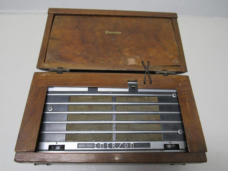 Puede incluir: Radio Emerson vintage con una caja de madera. La radio tiene una tapa con bisagras y una rejilla de altavoz con barras met&aacute;licas horizontales. El logotipo de Emerson es visible en la parte delantera. La caja es de madera y tiene un tono marr&oacute;n c&aacute;lido.