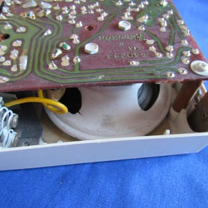 May include: Close-up of a vintage electronic component. A circuit board with green pathways and silver components sits above a white speaker. A yellow wire and a metal spring are also visible.