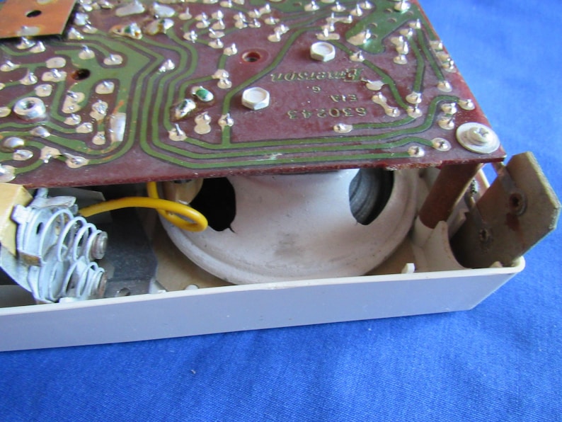 May include: Close-up of a vintage electronic component. A circuit board with green pathways and silver components sits above a white speaker. A yellow wire and a metal spring are also visible.