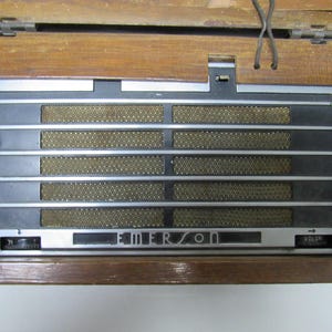Puede incluir: Radio Emerson vintage con una caja de madera. La parte frontal presenta una rejilla de altavoz con barras plateadas horizontales y un panel negro. La palabra "EMERSON" est&aacute; impresa en la parte delantera. La radio tiene una perilla de control de volumen.