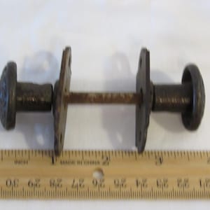 May include: An antique door latch with two decorative, dark metal knobs connected by a rusty rod. The knobs have a circular design. A wooden ruler with inch markings is in the foreground.