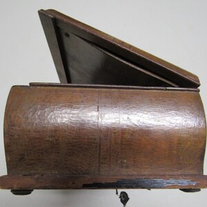 Puede incluir: Una caja de madera vintage con tapa con bisagras, parcialmente abierta. La caja tiene una superficie texturizada y tallada y un acabado marr&oacute;n oscuro. La tapa est&aacute; inclinada hacia arriba, revelando el interior. La caja descansa sobre peque&ntilde;os pies redondeados.