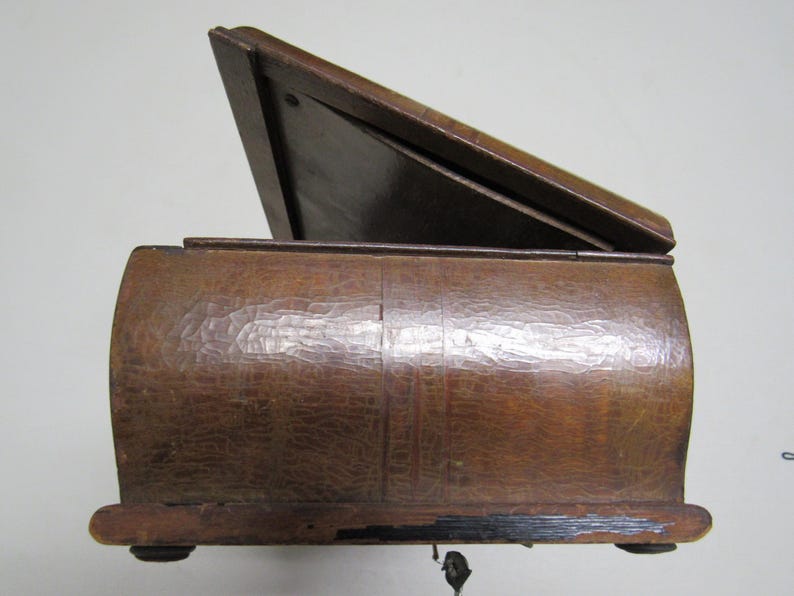 Puede incluir: Una caja de madera vintage con tapa con bisagras, parcialmente abierta. La caja tiene una superficie texturizada y tallada y un acabado marr&oacute;n oscuro. La tapa est&aacute; inclinada hacia arriba, revelando el interior. La caja descansa sobre peque&ntilde;os pies redondeados.