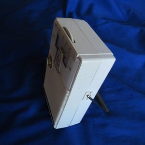 May include: A white vintage portable radio with a silver antenna and a tuning dial. The radio has a rectangular shape with a speaker grill on the front and a small dial on the side. The radio is set against a blue background.