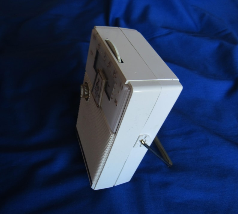 May include: A white vintage portable radio with a silver antenna and a tuning dial. The radio has a rectangular shape with a speaker grill on the front and a small dial on the side. The radio is set against a blue background.