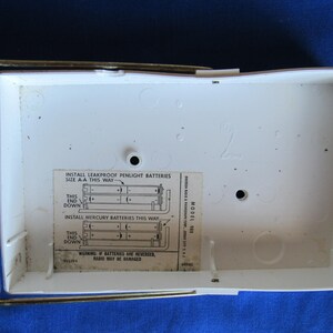 May include: A white radio battery compartment with a metal frame and a clear plastic cover. The interior has a label with instructions for installing batteries. The text on the label includes "INSTALL LEAKPROOF PENLIGHT BATTERIES" and "WARNING: IF BATTERIES ARE REVERSED, RADIO MAY BE DAMAGED."