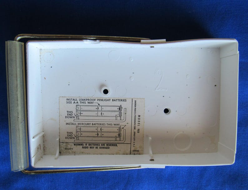 May include: A white radio battery compartment with a metal frame and a clear plastic cover. The interior has a label with instructions for installing batteries. The text on the label includes "INSTALL LEAKPROOF PENLIGHT BATTERIES" and "WARNING: IF BATTERIES ARE REVERSED, RADIO MAY BE DAMAGED."