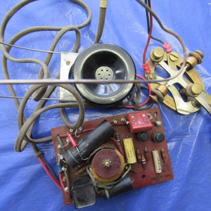 May include: A vintage telephone handset with a black receiver, a brown cord, and a circuit board with red, black, and green wires. The circuit board has a red capacitor, a black transformer, and a silver switch with a black knob. The text on the circuit board reads "2M1002 0444".