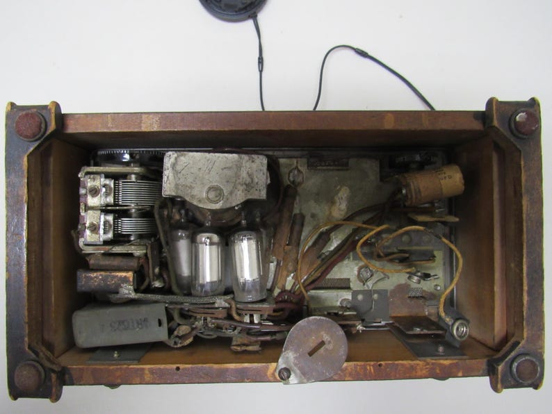 Puede incluir: Los componentes internos de una radio antigua son visibles dentro de un marco de madera. El interior presenta v&aacute;lvulas de vac&iacute;o, condensadores y cableado, mostrando la intrincada tecnolog&iacute;a del dise&ntilde;o de radio antiguo. El marco de madera tiene herrajes envejecidos.