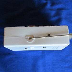 May include: A white, rectangular vintage music box with a silver metal crank and a small speaker. The top edge has a ridged design. The music box is set against a blue background.