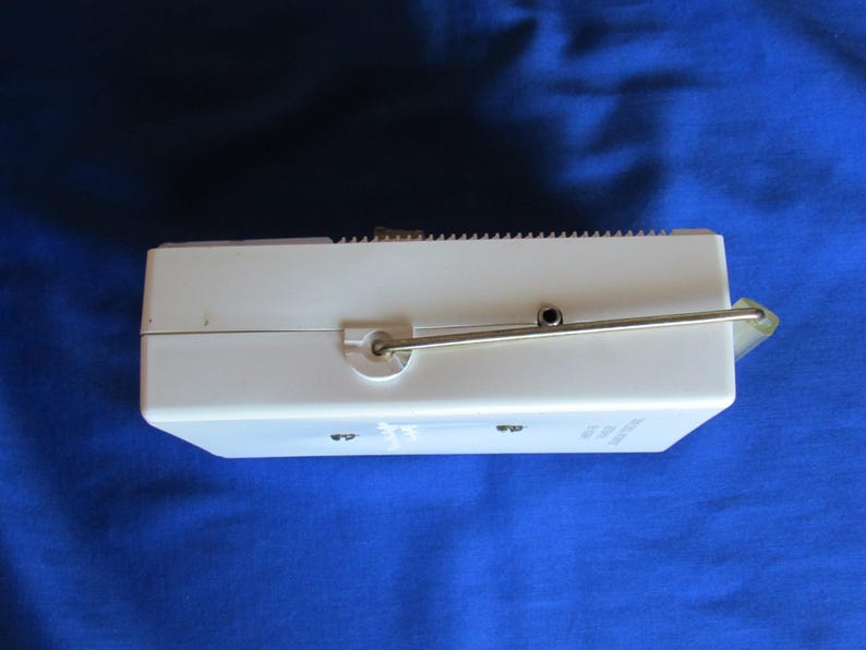 May include: A white, rectangular vintage music box with a silver metal crank and a small speaker. The top edge has a ridged design. The music box is set against a blue background.