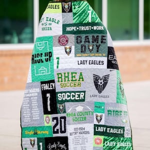 Pu&ograve; includere: Una trapunta ricordo realizzata con varie magliette sportive verdi, bianche, nere e grigie con grafiche a tema calcistico, nomi di squadre come Lady Eagles, numeri di maglia e testi come Hope Trust Work e History Makers, drappeggiata su una persona che indossa un tocco di laurea verde.