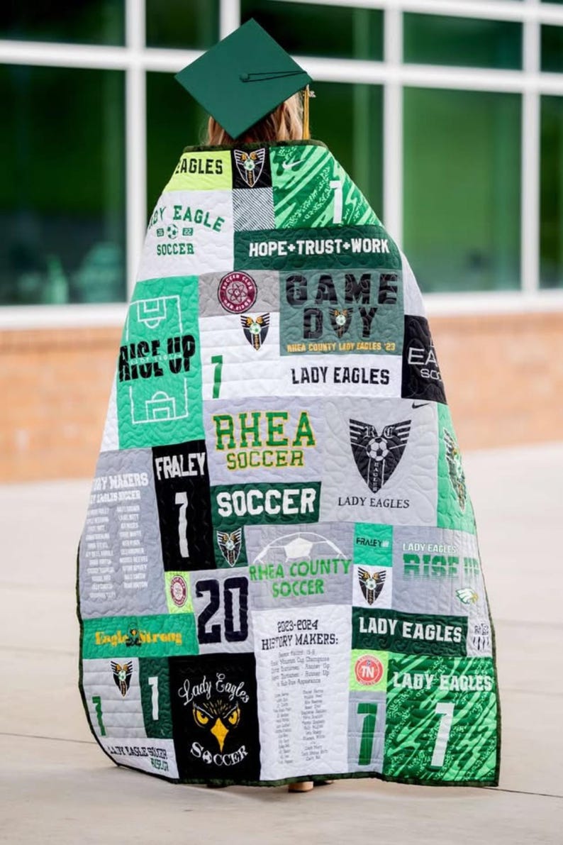 Pu&ograve; includere: Una trapunta ricordo realizzata con varie magliette sportive verdi, bianche, nere e grigie con grafiche a tema calcistico, nomi di squadre come Lady Eagles, numeri di maglia e testi come Hope Trust Work e History Makers, drappeggiata su una persona che indossa un tocco di laurea verde.