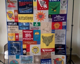 May include: A rectangular quilt constructed from various colorful t-shirt panels featuring diverse graphic prints, including text such as Kittatinny, Yorktown, Staff, Troop 171, and Conclave, alongside illustrations of wolves, ships, and eagles.