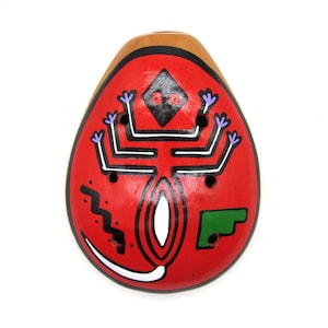 May include: A red clay ocarina with a black and white design of a stylized figure with purple flowers. The ocarina has six finger holes and a green accent.