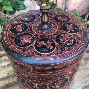 以下が含まれることがあります： 動物と花のモチーフが複雑に手描きされた、黒と赤の円筒形のランプベース。ランプベースは木でできており、真鍮製のランプソケットが付いています。