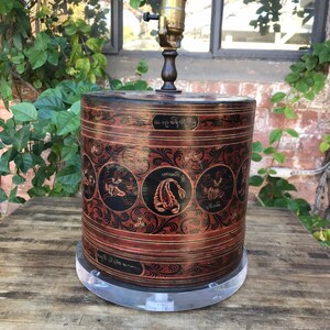 以下が含まれることがあります： サソリやその他のシンボルが描かれた黒と赤のペイントデザインが施された木製ランプベース。ベースは円筒形で、透明なアクリルベースの上に置かれています。