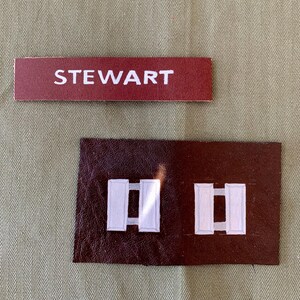 Puede incluir: Una etiqueta de nombre granate con el nombre "STEWART" en letras blancas. Debajo hay un parche marrón oscuro con dos barras blancas, probablemente insignias militares. Los artículos están sobre un fondo verde oliva texturizado.