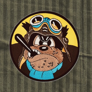 Puede incluir: Parche bordado circular que representa un perro de dibujos animados con un casco y gafas de aviador marrones. El perro sostiene un objeto negro, sobre un círculo amarillo y un fondo a rayas verdes. El diseño tiene una estética vintage de inspiración militar.